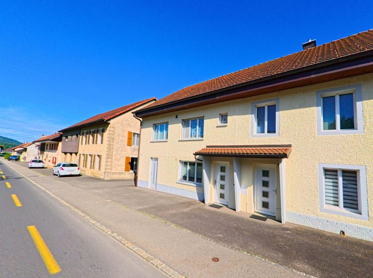 In Develier (2802, Kanton Jura) sind moderne und traditionelle Häuser entlang der Straße zu sehen, mit Autos, die am Straßenrand geparkt sind.