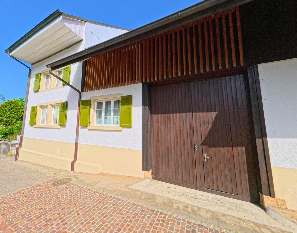 Ein traditionelles Haus in Diegten (4457, Kanton Baselland) mit grünen Fensterlaeden und grossem Holztor liegt an einer gepflasterten Strasse unter blauem Himmel.