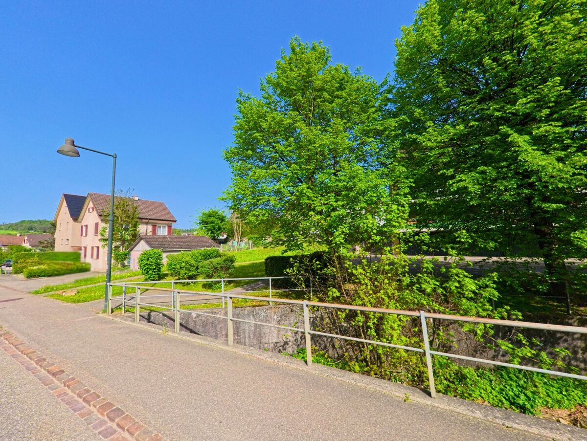 Ein gepflasterter Gehweg mit Geländer verläuft in Diegten (4457, Kanton Baselland) entlang eines kleinen Bachs, flankiert von Wohnhaeusern und dichtem Gruen.