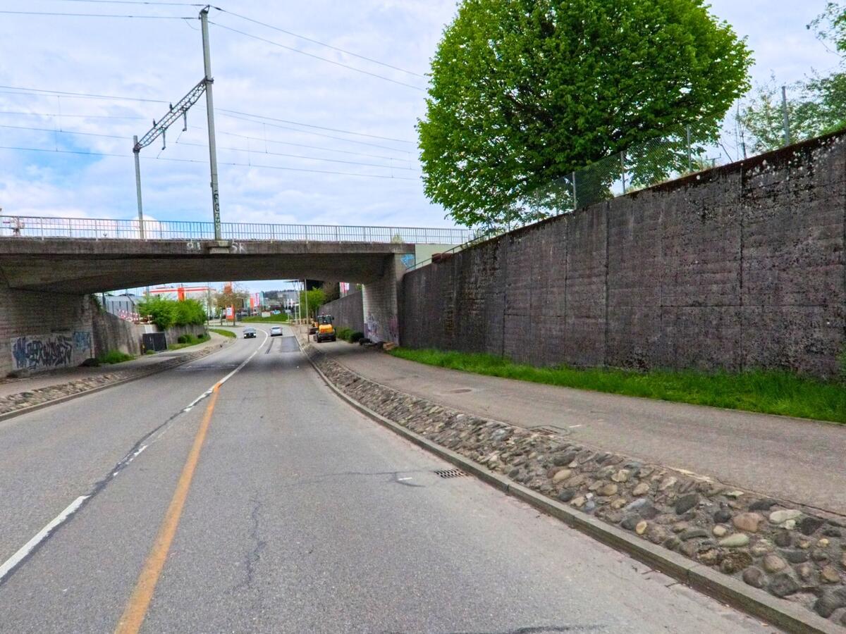 Eine Strasse in Dielsdorf, PLZ 8157 im Kanton Zürich, führt unter einer Eisenbahnbrücke hindurch, flankiert von hohen Betonmauern und einem Gehweg.
