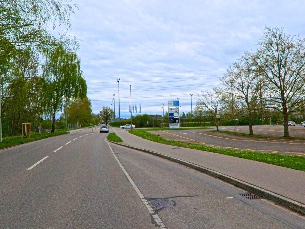  In Dielsdorf, PLZ 8157 im Kanton Zürich, verläuft eine Strasse entlang eines grossen Parkplatzes nahe eines Sport- und Gewerbegebiets.