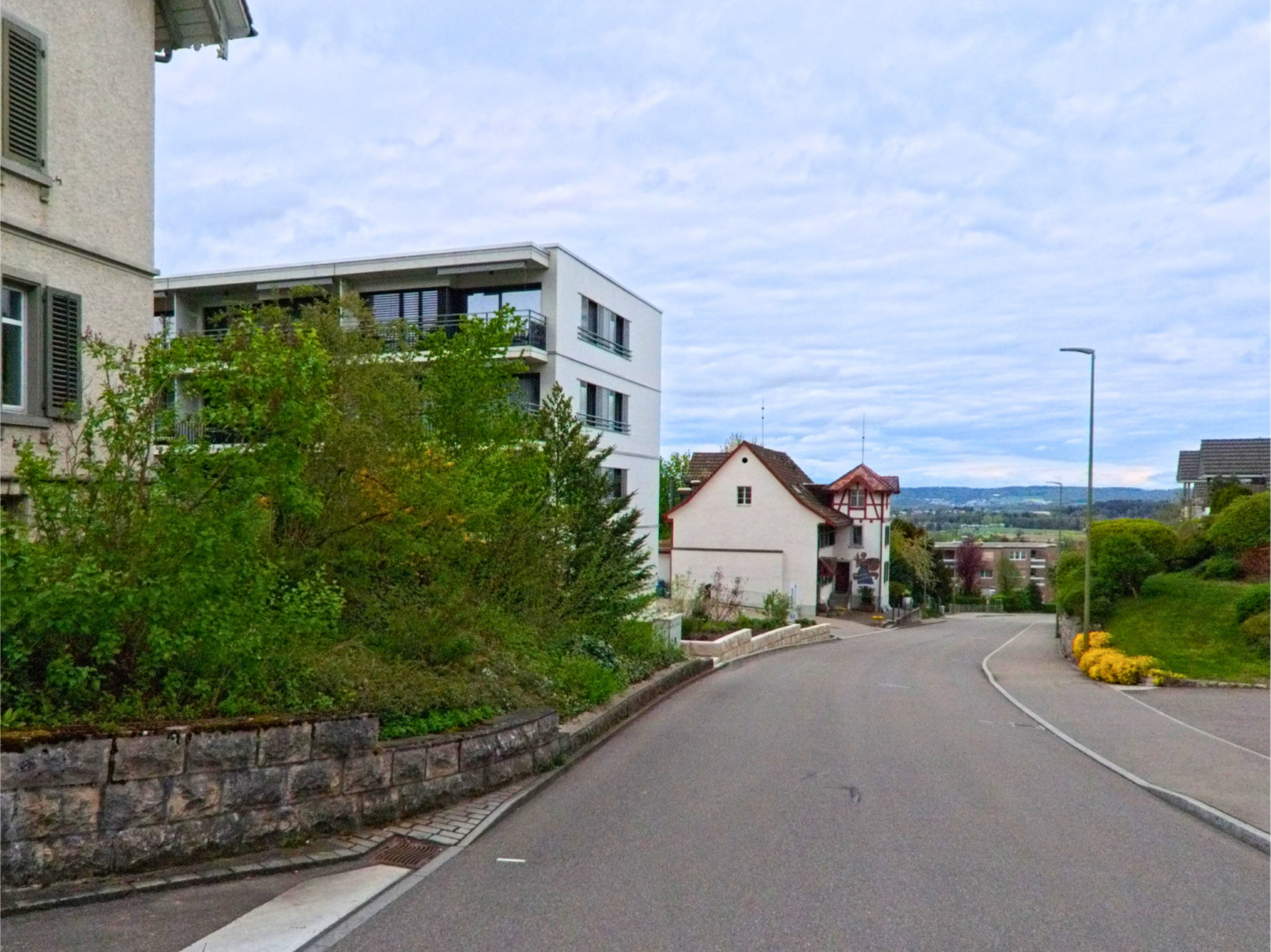 In Dielsdorf, PLZ 8157 im Kanton Zürich, führt eine leicht abschüssige Strasse vorbei an modernen und traditionellen Wohnhäusern mit Blick ins Umland.