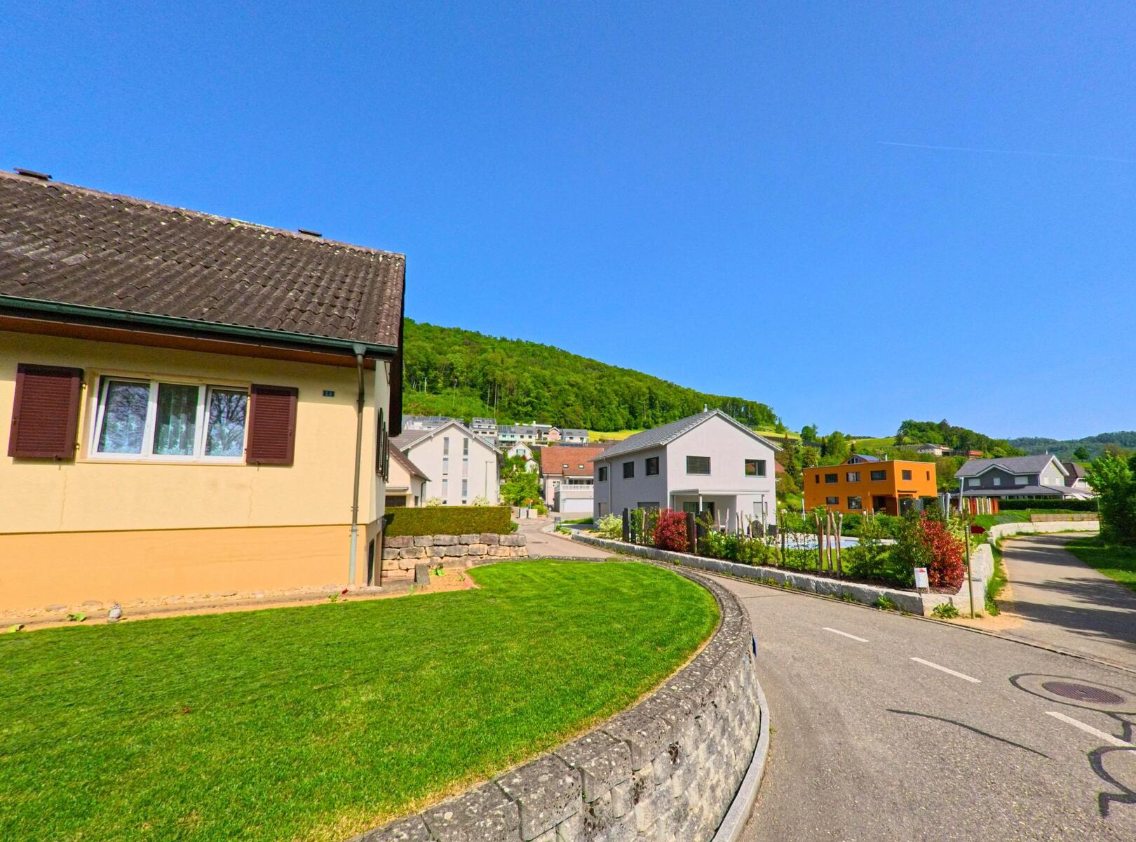 Diepflingen (4442, Kanton Baselland) führt eine ruhige Strasse entlang moderner Häuser, umgeben von gepflegten Gärten und grünen Hügeln im Hintergrund.