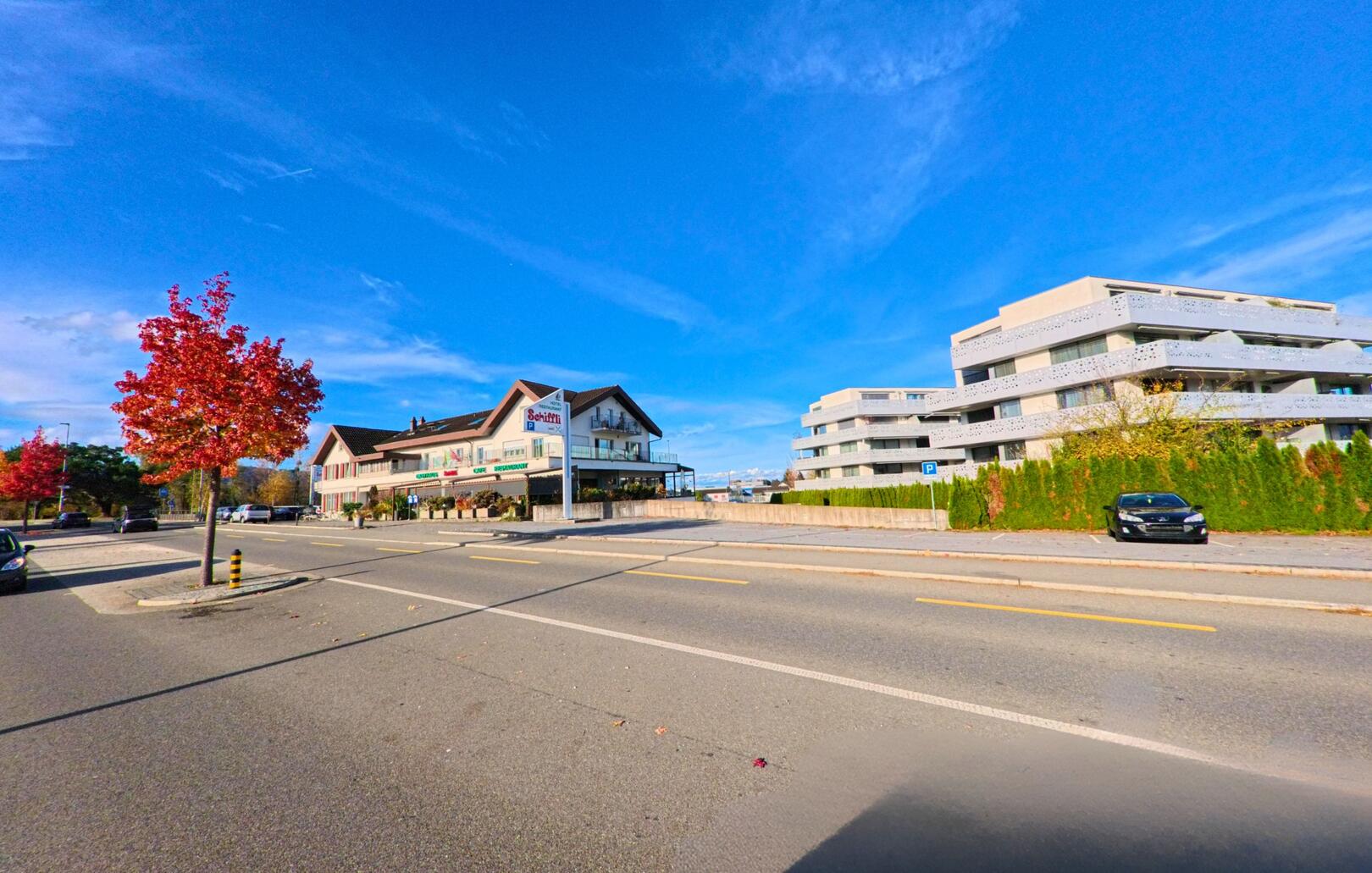 Vor blauem Himmel zeigt sich in Diepoldsau (9444, Kanton St. Gallen) eine breite Strasse mit einem Wohnhaus links und modernen Gebäuden rechts.
