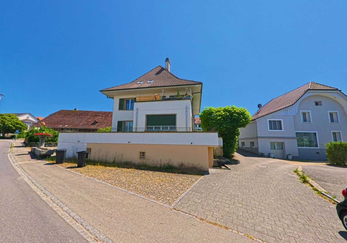 Ein Mehrfamilienhaus mit markantem Walmdach steht an einer leichten Kurve in Diessbach bei Büren (3264, Kanton Bern), flankiert von einem Brunnen und gepflegten Nachbarhäusern.