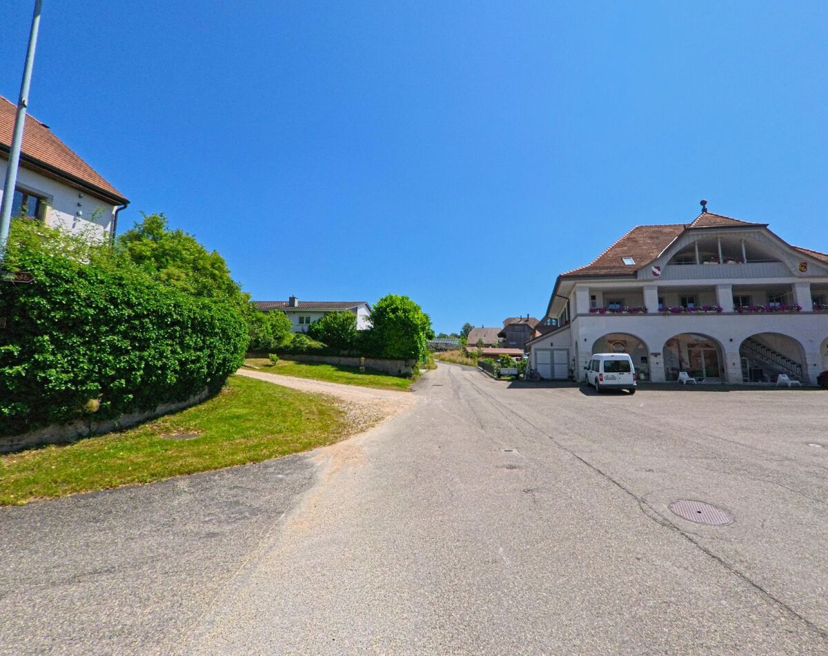 In Diessbach bei Büren (3264, Kanton Bern) führt eine ruhige Straße mit angrenzendem Kiesweg bergauf, vorbei an einem Gebäude mit Arkaden und Balkonblumen.