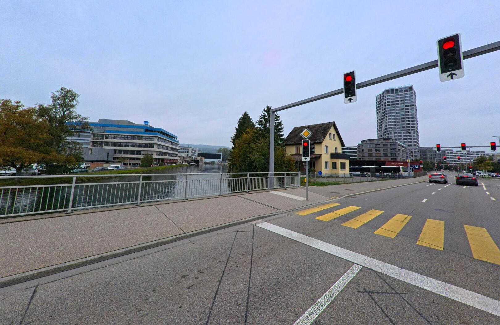 An der Strasse von Dietikon (8953, Kanton Zürich) ueberblickt die Aufnahme eine Bruecke am Fluss, flankiert von modernen Gebaeuden und einem gelben Wohnhaus bei Rotlicht.