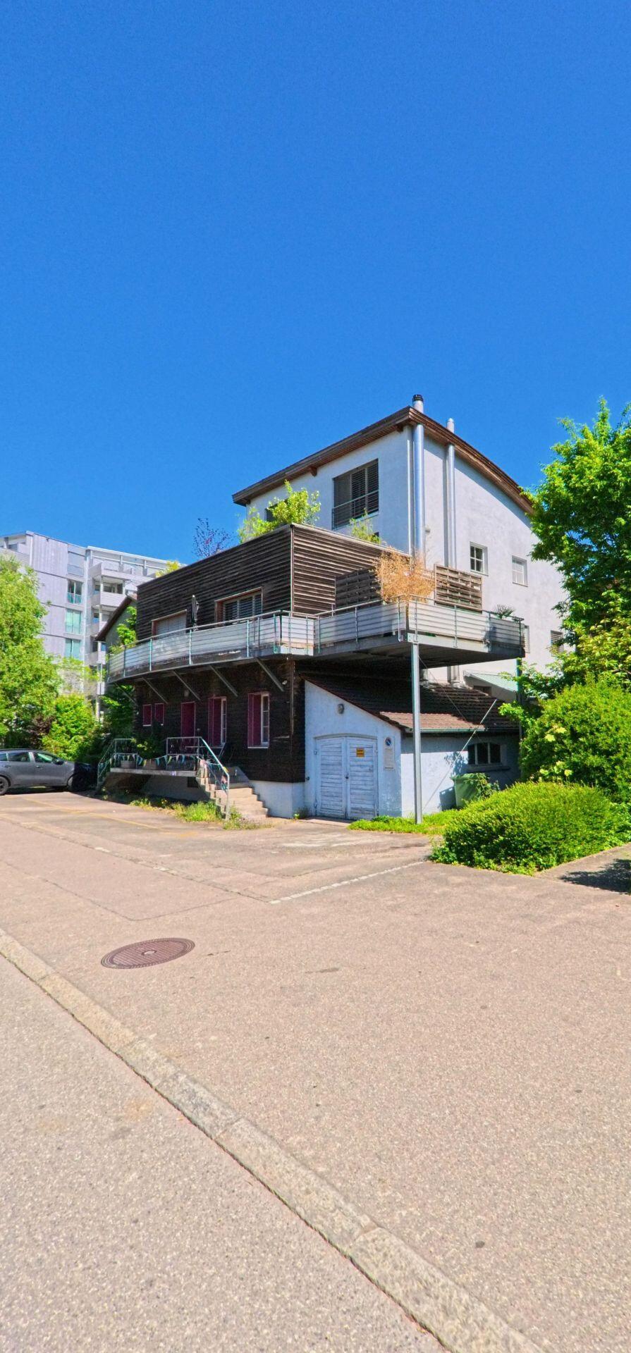 In Dietikon (8953, Kanton Zürich) steht ein mehrgeschossiges Haus mit grosser Terrasse und Holzverkleidung, umgeben von Gruen und angrenzenden modernen Wohnbauten.
