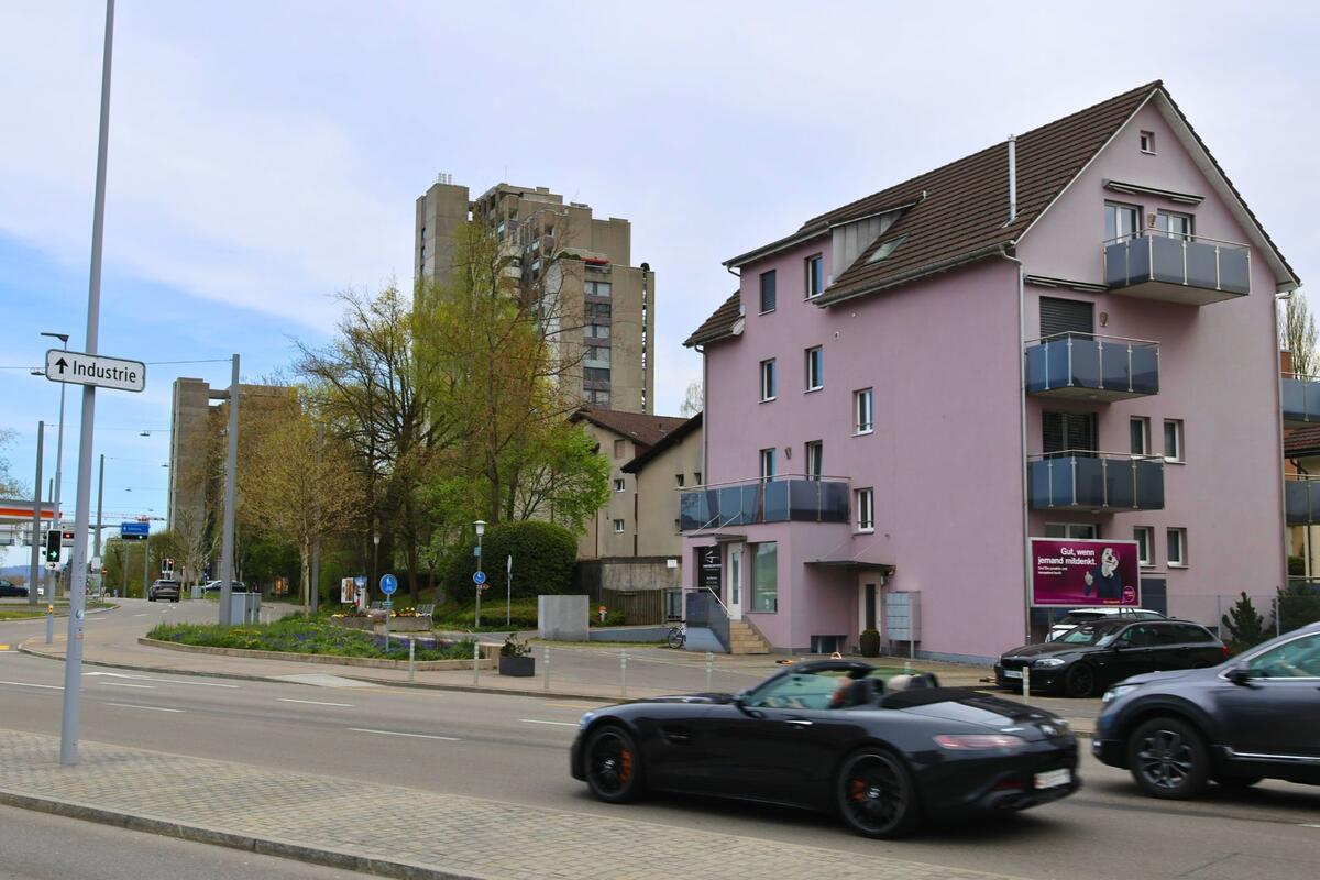  In Dietikon, PLZ 8953 im Kanton Zürich, verläuft eine vielbefahrene Strasse mit Hochhäusern im Hintergrund und einem auffällig rosafarbenen Wohnhaus im Vordergrund.
