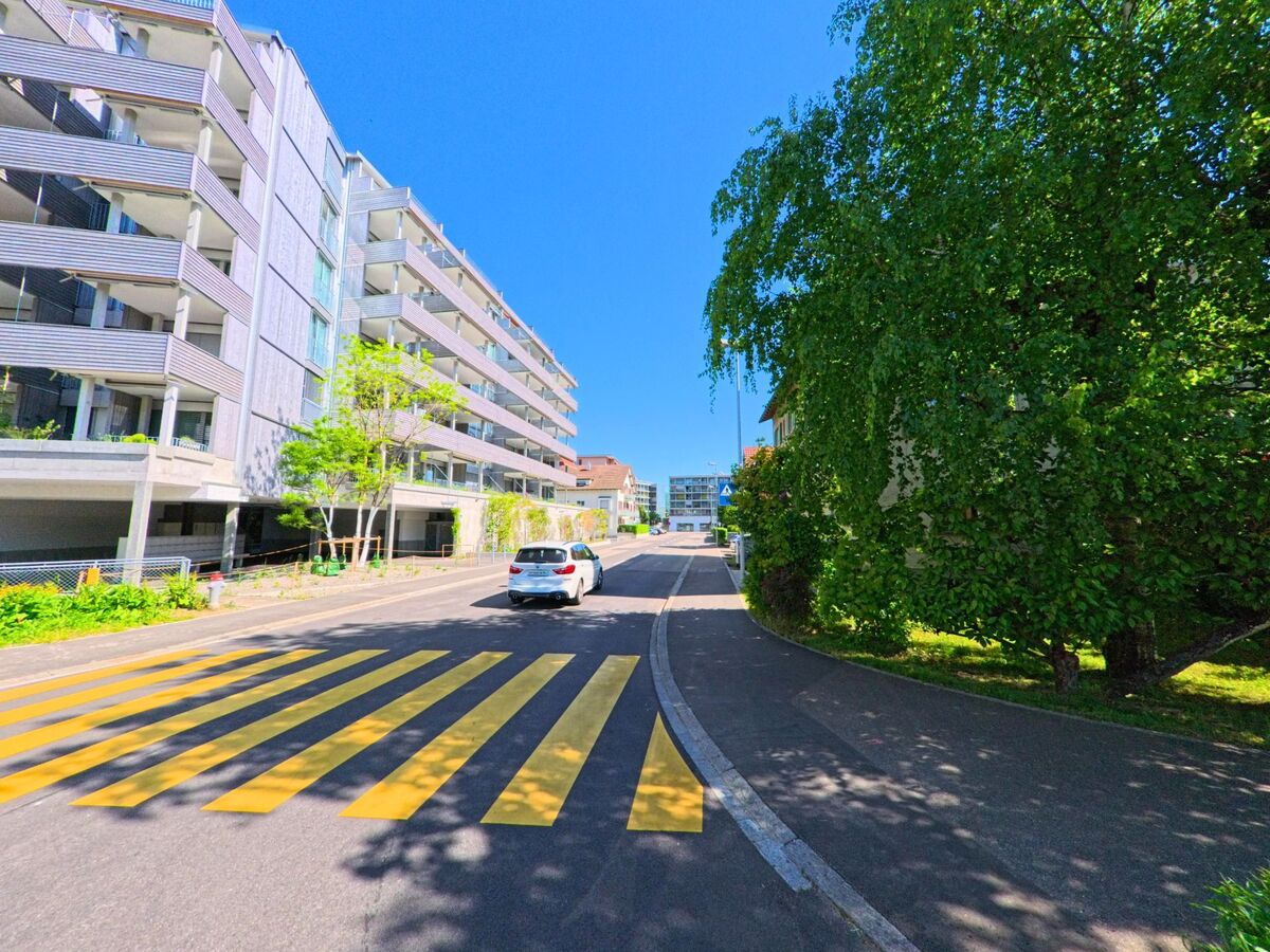 In Dietikon im Kanton Zürich, PLZ 8953, verläuft eine Quartierstrasse mit modernen Wohnbauten und einem markierten Fussgängerstreifen im Sonnenschein.