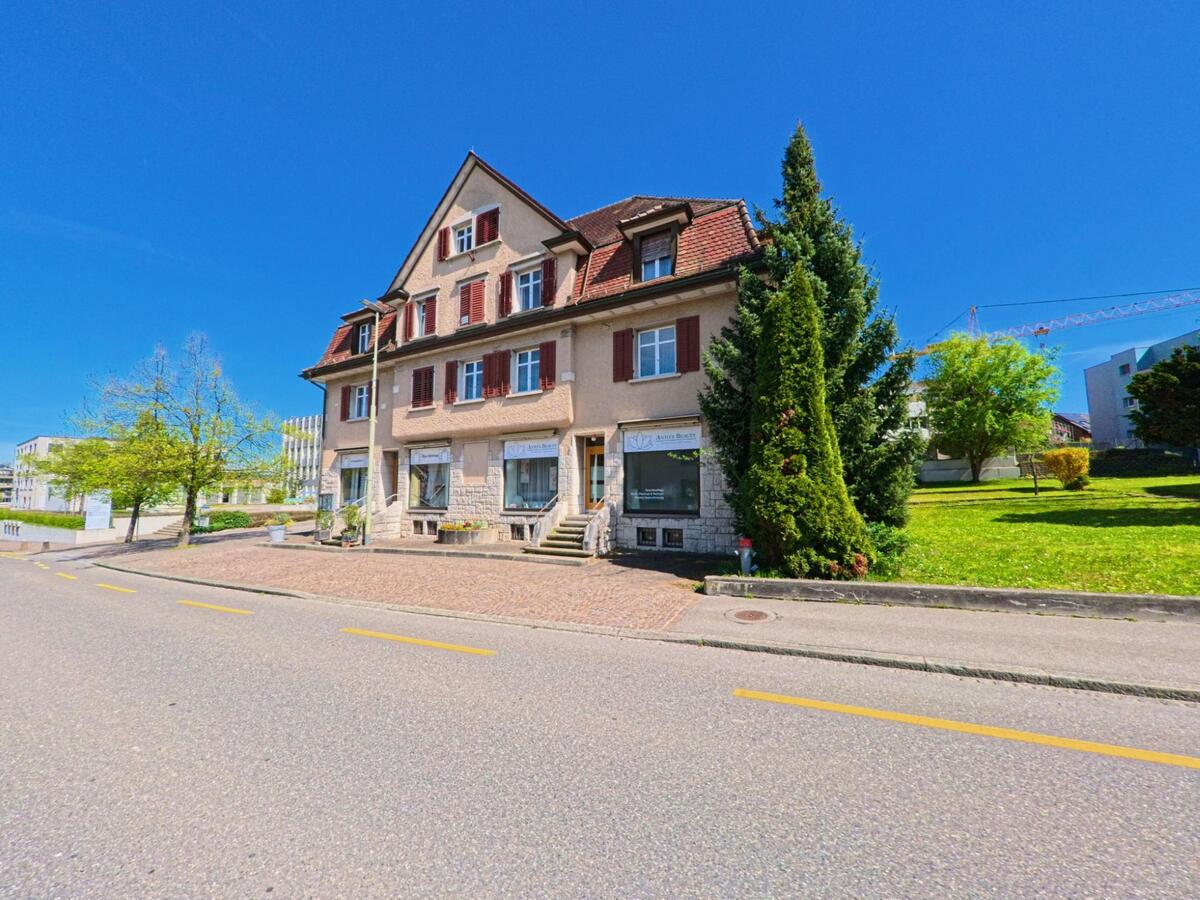 In Dietlikon im Kanton Zürich, PLZ 8305, befindet sich ein historisches Gebäude mit Schaufenstern entlang einer ruhigen Strasse, umgeben von gepflegtem Grün.