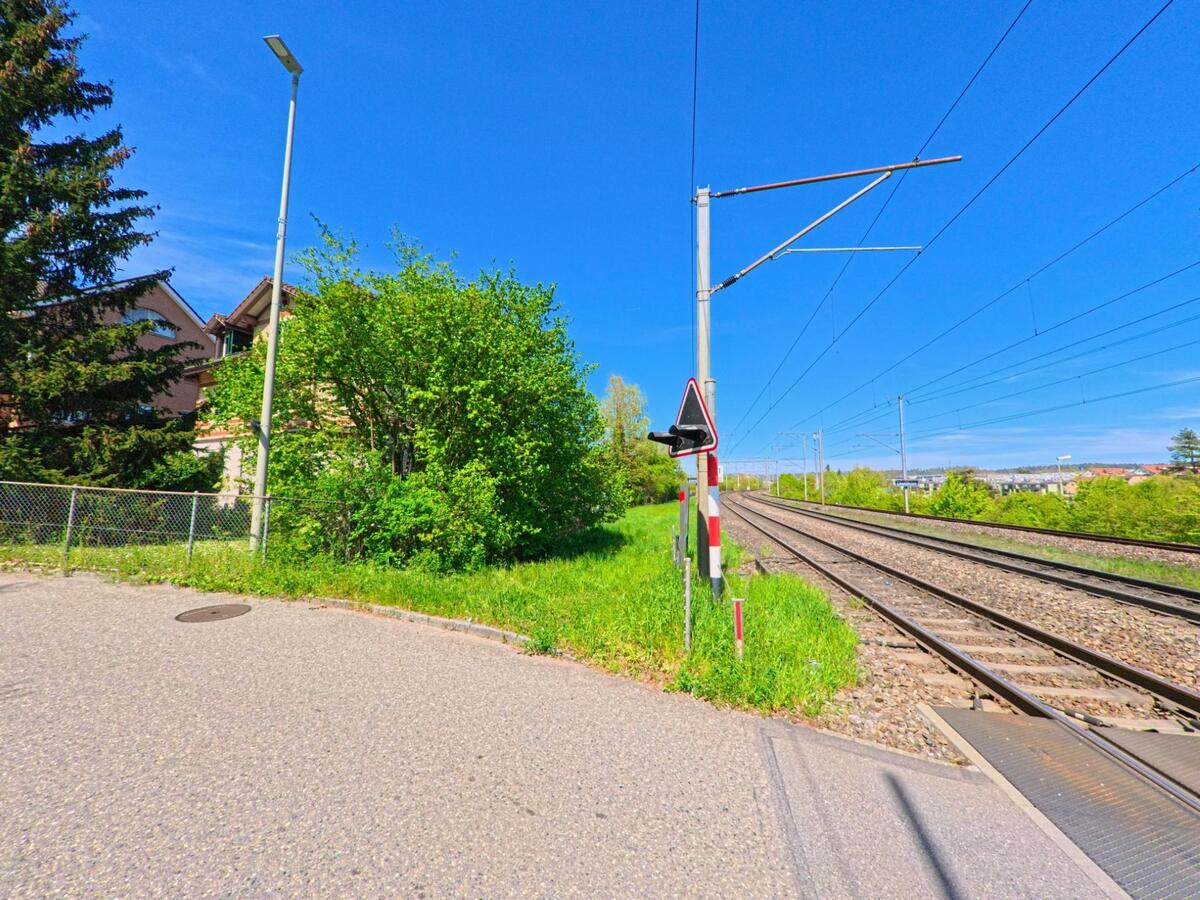 In Dietlikon im Kanton Zürich, PLZ 8305, verläuft eine Bahnstrecke entlang eines Fusswegs mit Warnschild und angrenzender Wohnbebauung im Hintergrund.