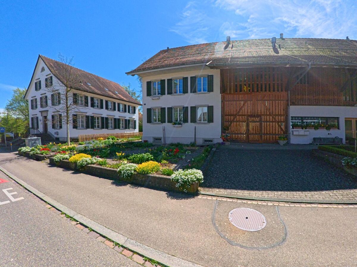 In Dietlikon, PLZ 8305 im Kanton Zürich, stehen ein historisches Bauernhaus mit grossem Holztor und ein angrenzendes Wohnhaus hinter einem gepflegten Blumengarten.