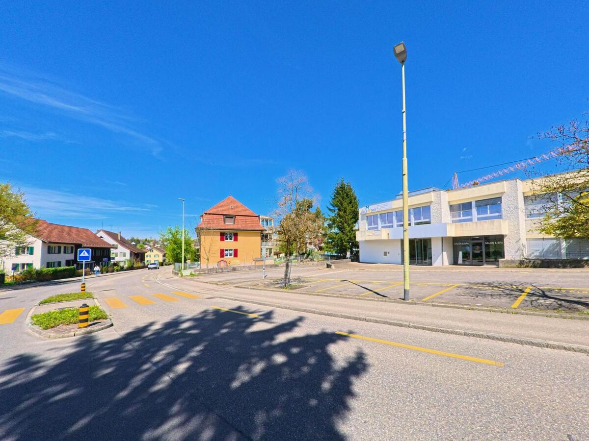 In Dietlikon im Kanton Zürich, PLZ 8305, trifft eine leicht gebogene Strasse auf ein kleines Geschäftsgebäude und Wohnhäuser mit markierten Fussgängerstreifen.