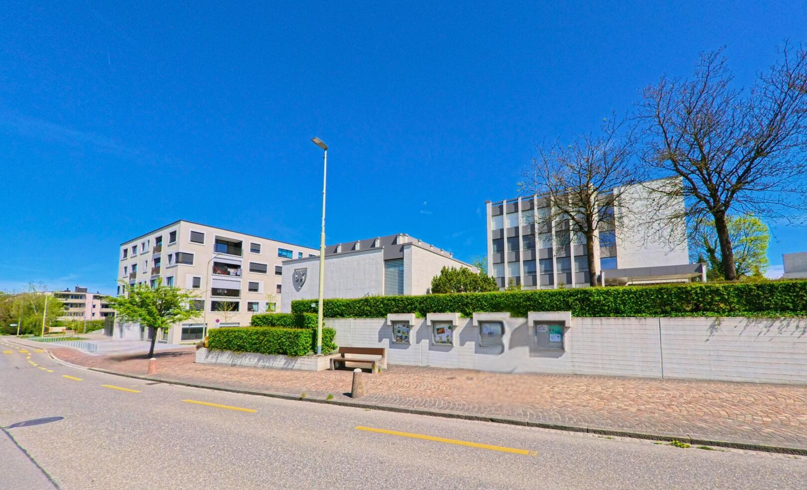 In Dietlikon im Kanton Zürich, PLZ 8305, verläuft eine ruhige Strasse entlang moderner Wohn- und Verwaltungsgebäude mit gepflegtem Vorgarten und Anschlagtafeln.