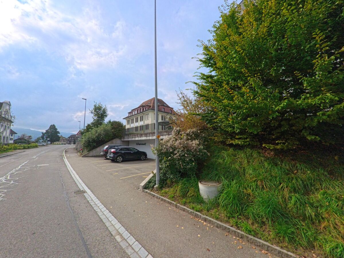 Entlang der leicht ansteigenden Strasse in Dietwil (6042, Kanton Aargau) liegen Parkplätze und das markante Gebäude des Obermatt mit Blick auf das Dorfzentrum.