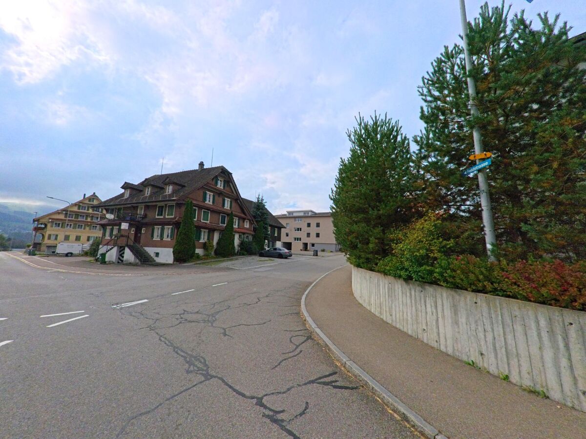 An einer Kreuzung in Dietwil (6042, Kanton Aargau) stehen traditionelle Holzhäuser mit grünen Fensterläden neben neueren Gebäuden und einer leicht ansteigenden Strasse.