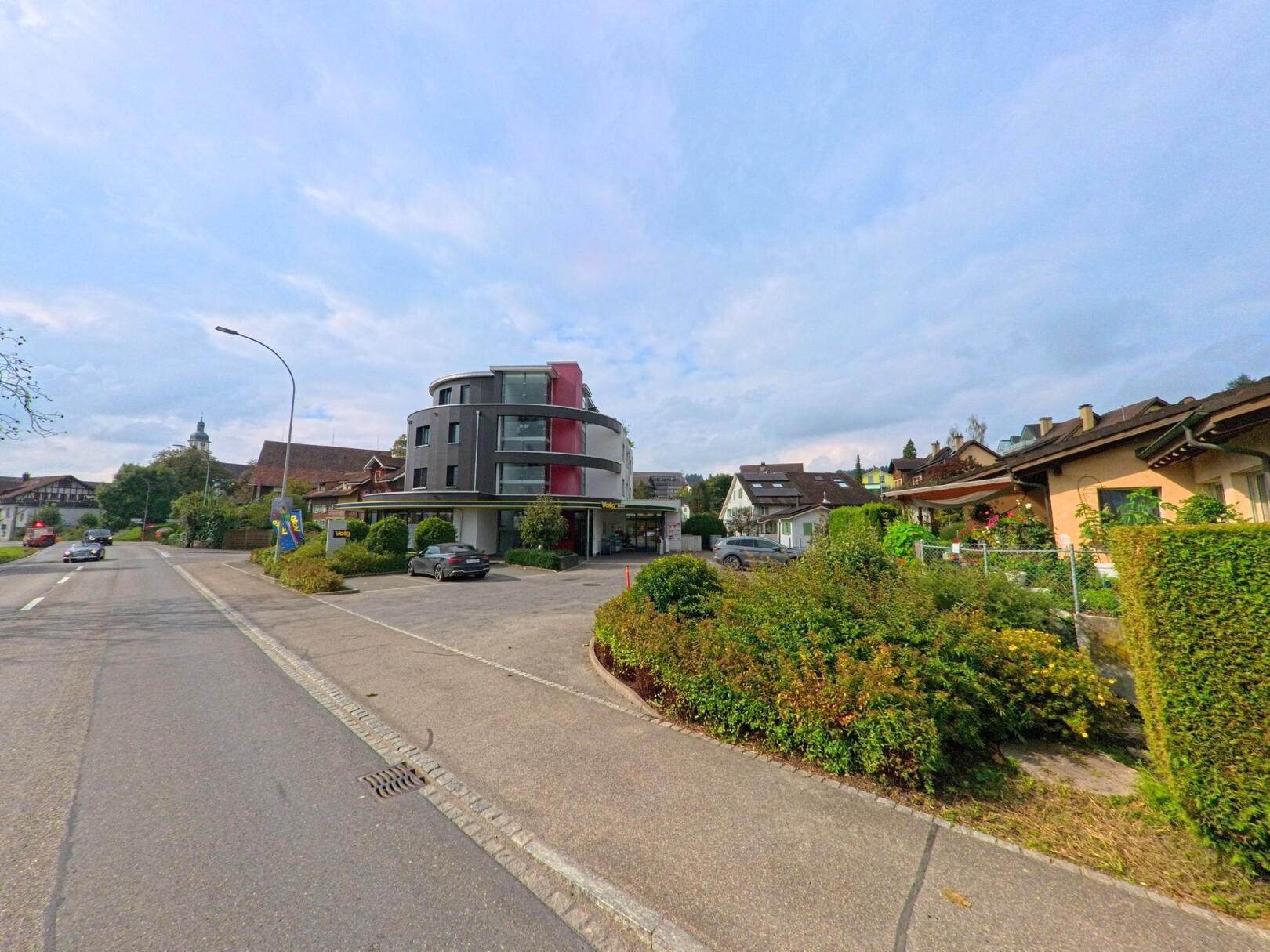 In Dietwil (6042, Kanton Aargau) steht ein modernes rundes Wohn- und Geschäftsgebäude entlang der Hauptstrasse, umgeben von gepflegten Gärten und Einfamilienhäusern.