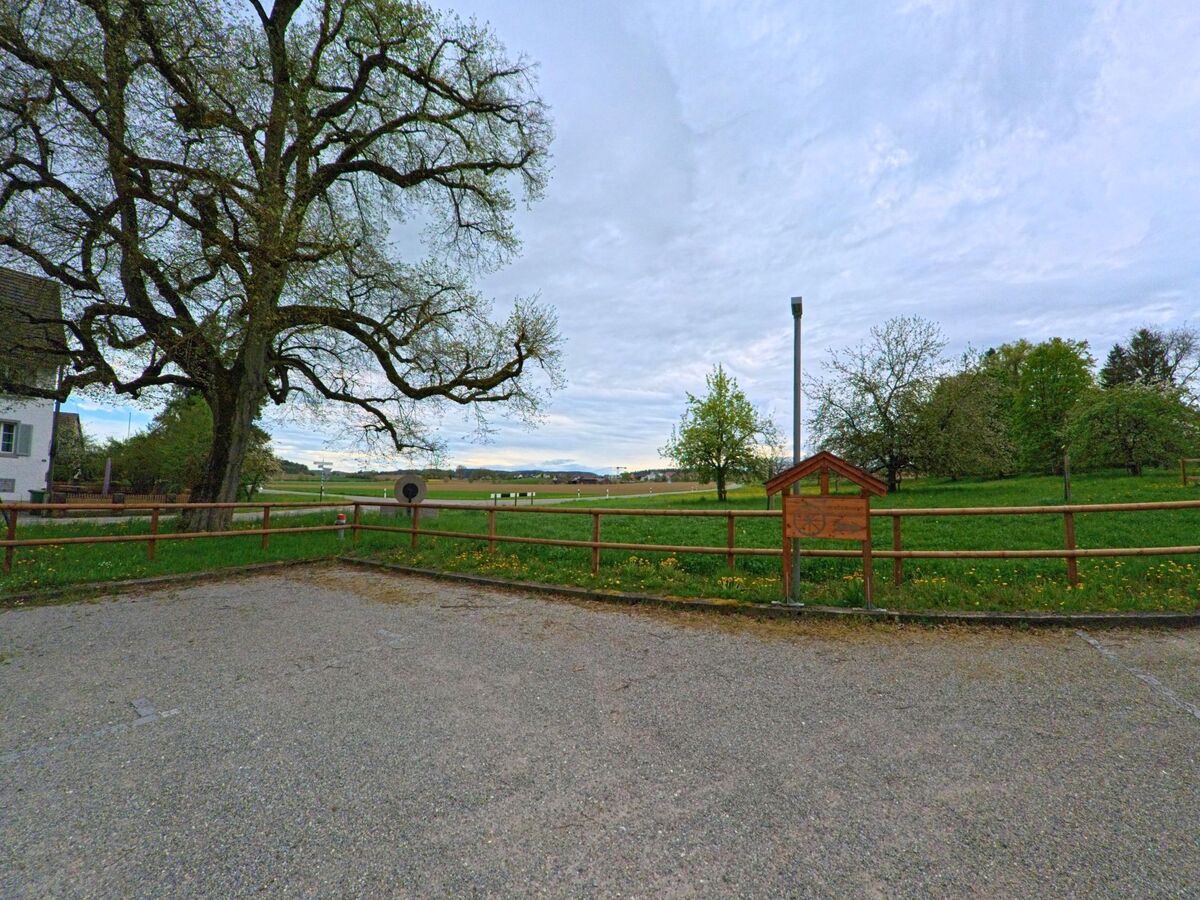In Dinhard im Kanton Zürich, PLZ 8474, bietet ein kleiner Rastplatz mit Holztafel und umzäunter Wiese einen weiten Blick über die Felder bis zum Horizont.
