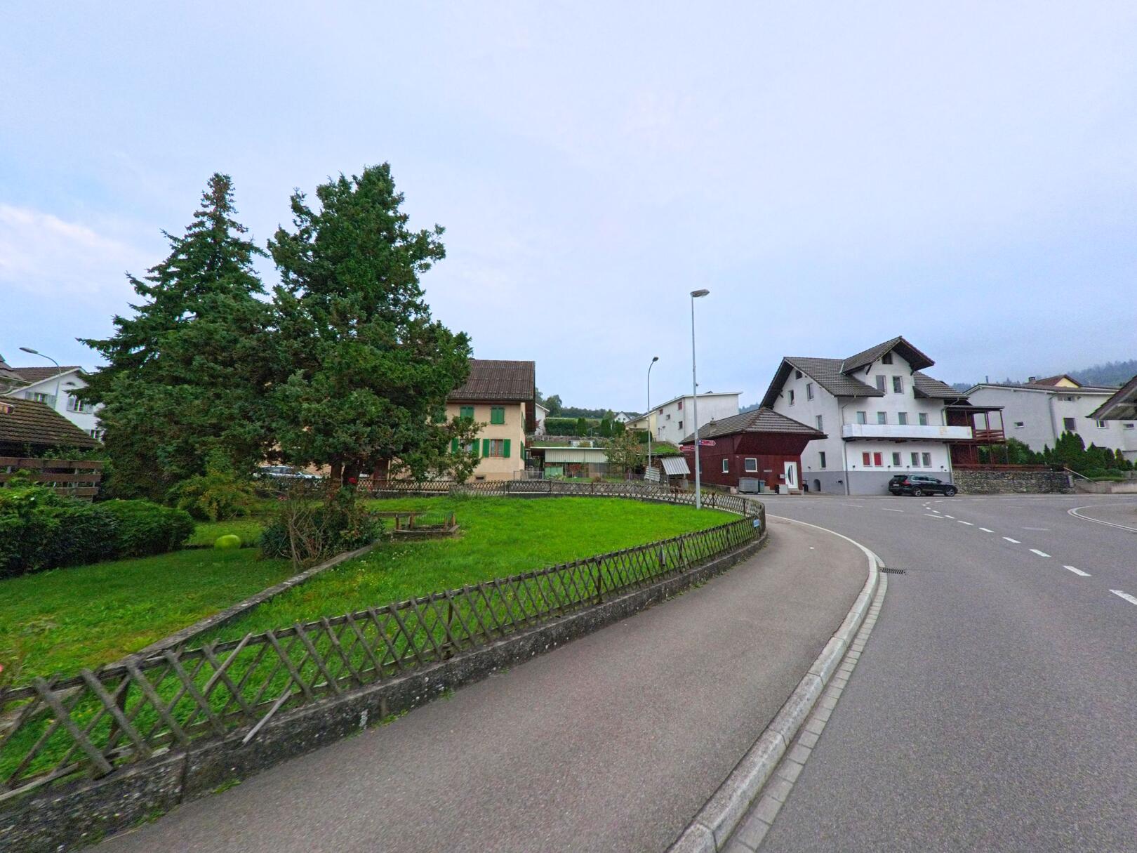 An einer sanft geschwungenen Strasse in Dintikon (5606, Kanton Aargau) stehen Einfamilienhäuser mit gepflegten Gärten und hohen Bäumen.