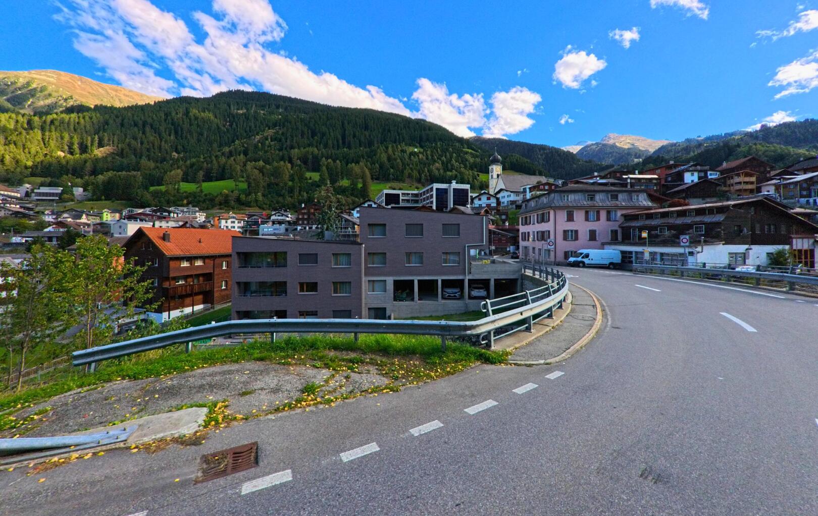 Eine kurvige Strasse fuehrt durch Disentis/Muster (7180, Kanton Graubuenden) mit Blick auf die Kirche und die umgebenden Wohnhaeuser vor bewaldeten Berghaengen.