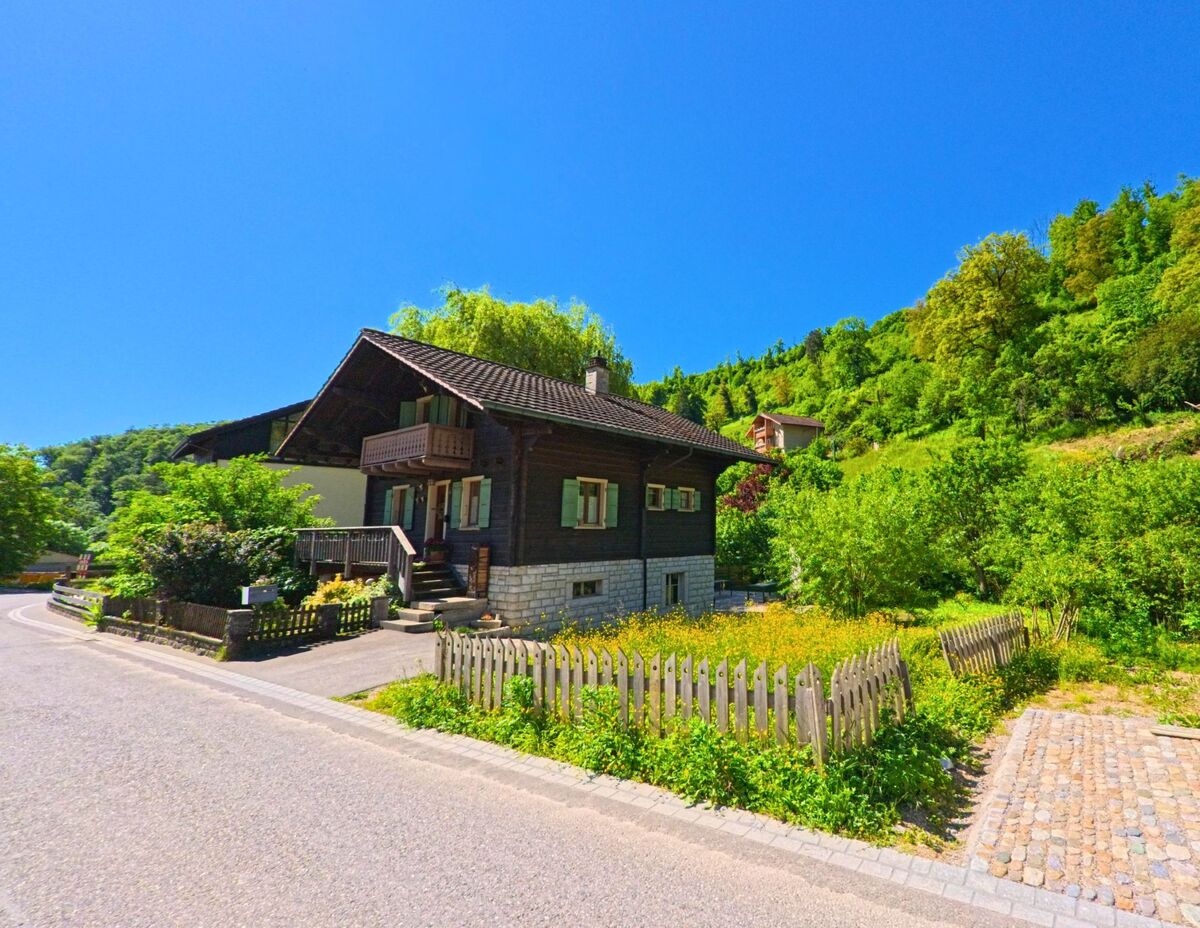 Ein Chaletstil-Haus mit gruener Fensterlaeden steht in Dittingen (4243, Kanton Baselland) am Waldrand an einer leicht geschwungenen Strasse mit Naturgarten.