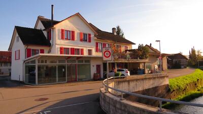 Die Dorf-Schmiede Bed&Breakfast in Müllheim (8555, Kanton Thurgau) ist ein traditionelles Gebaeude mit roten Fensterlaeden am Strassenrand nahe eines Baches.