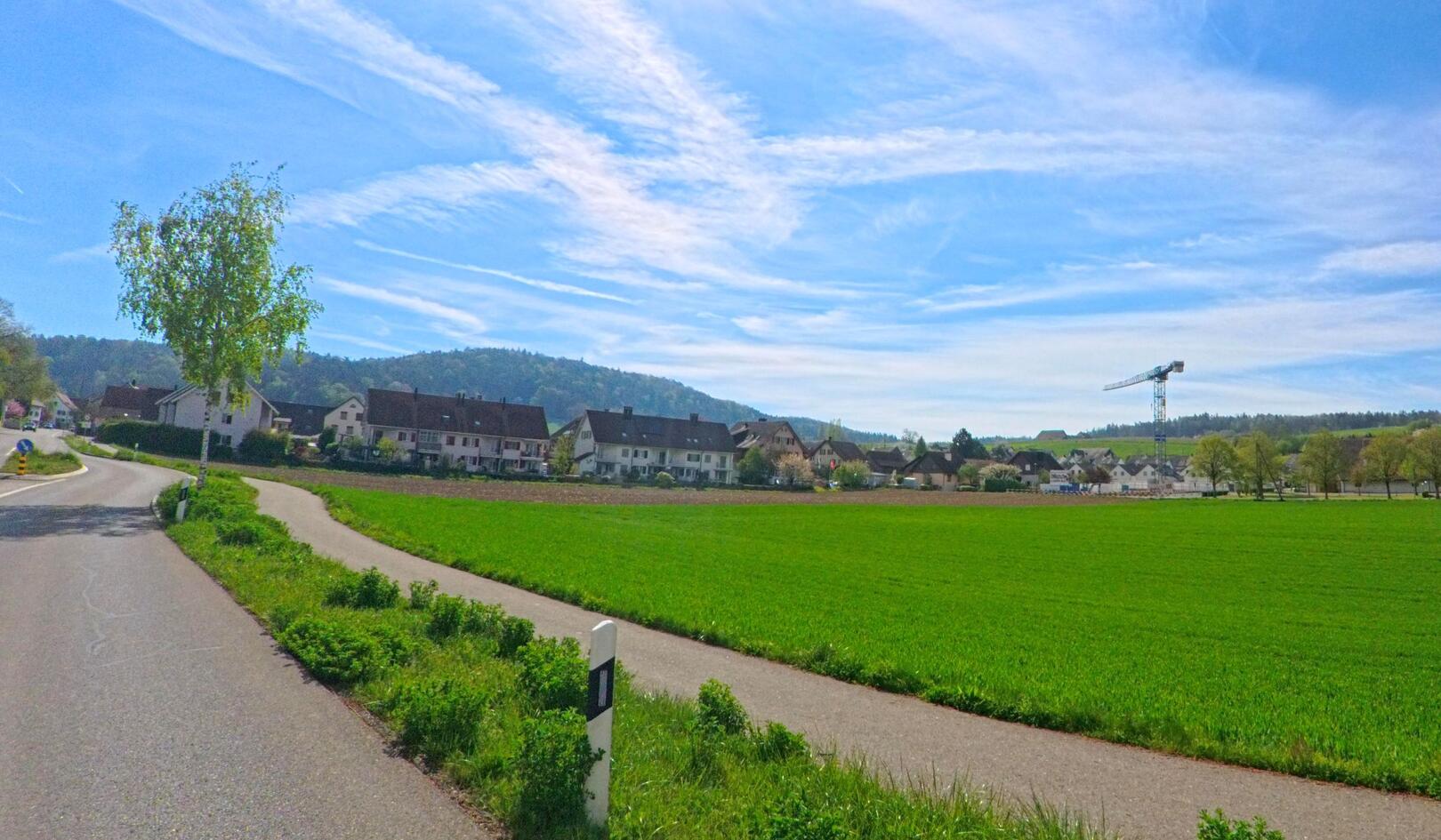 In [ORT] mit der Postleitzahl [PLZ] im [KANTON] verläuft ein geteilter Weg entlang grüner Felder, während im Hintergrund Wohnhäuser und ein Baukran das Dorfbild prägen.