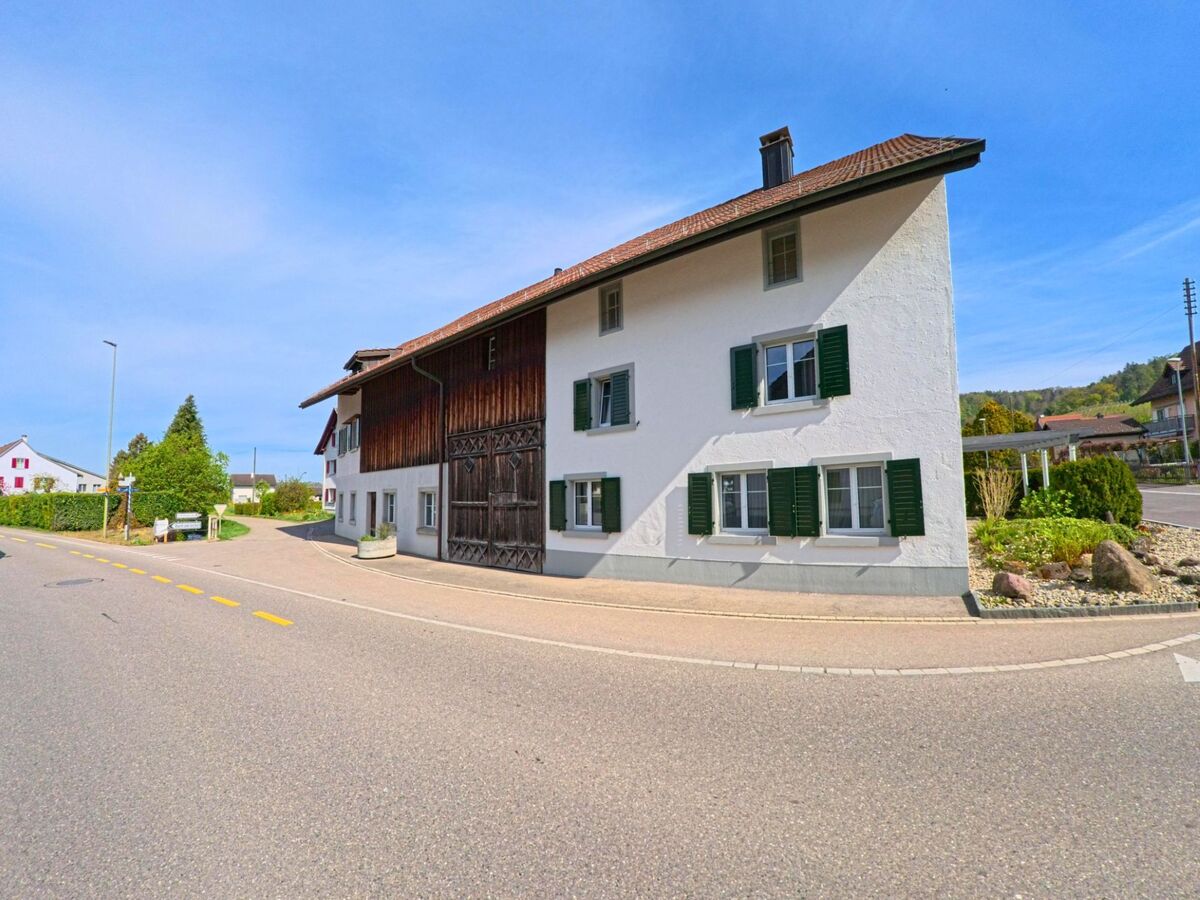 Ein Wohnhaus mit grossem Holztor und grünen Fensterläden steht in [ORT], PLZ [PLZ], im [KANTON] an einer ruhigen Strassenkreuzung mit gepflegter Umgebung.