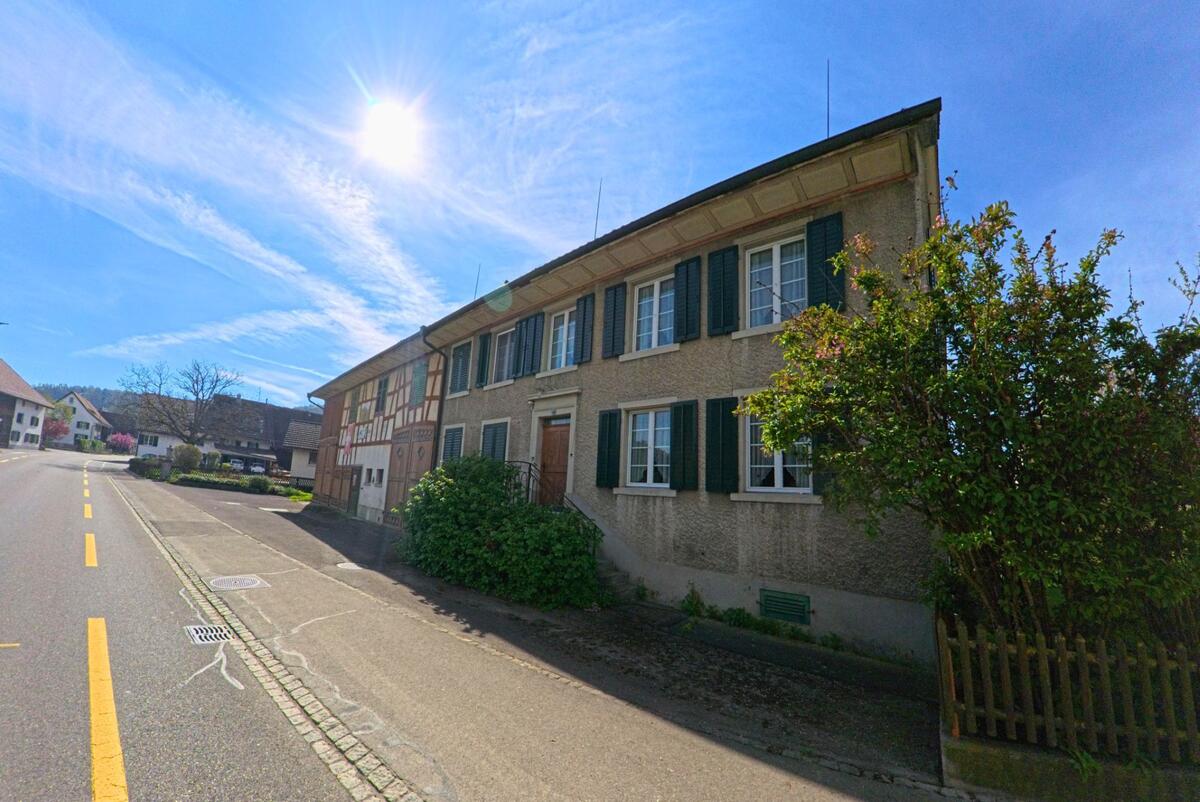 In [ORT], PLZ [PLZ], im [KANTON] reiht sich ein traditionelles Haus mit grünen Fensterläden neben einem Fachwerkhaus an einer sonnigen Strasse.