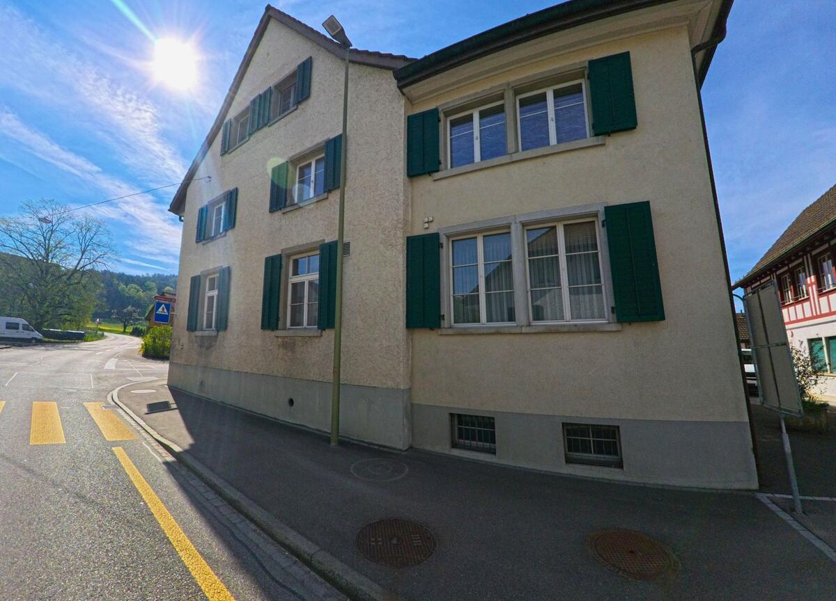Ein Fussgängerstreifen führt in [ORT], PLZ [PLZ], im [KANTON] an einem alten Wohnhaus mit grünen Fensterläden vorbei, während die Sonne tief am Himmel steht.
