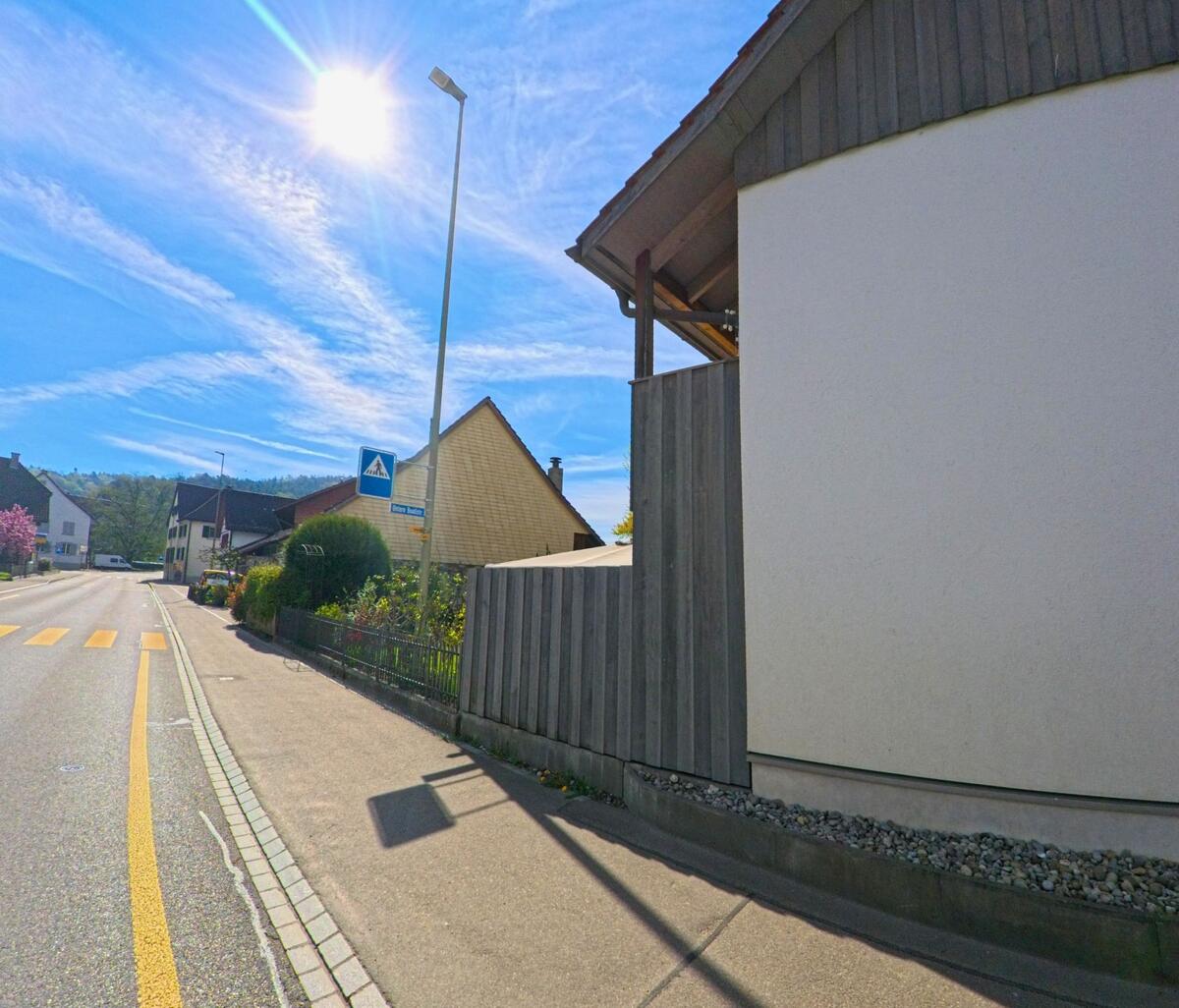An einer verkehrsberuhigten Strasse in [ORT], PLZ [PLZ], im [KANTON] verläuft ein Fussgängerstreifen neben modernen und traditionellen Häusern unter klarem Himmel.