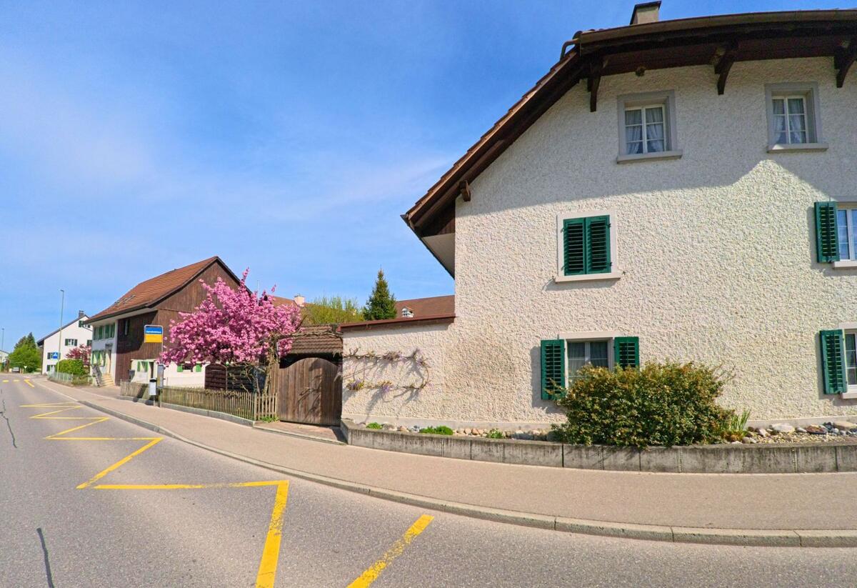In [ORT], PLZ [PLZ], im [KANTON] liegt eine Haltestelle an einer Strasse mit traditionellen Wohnhäusern, blühenden Bäumen und grünen Fensterläden.
