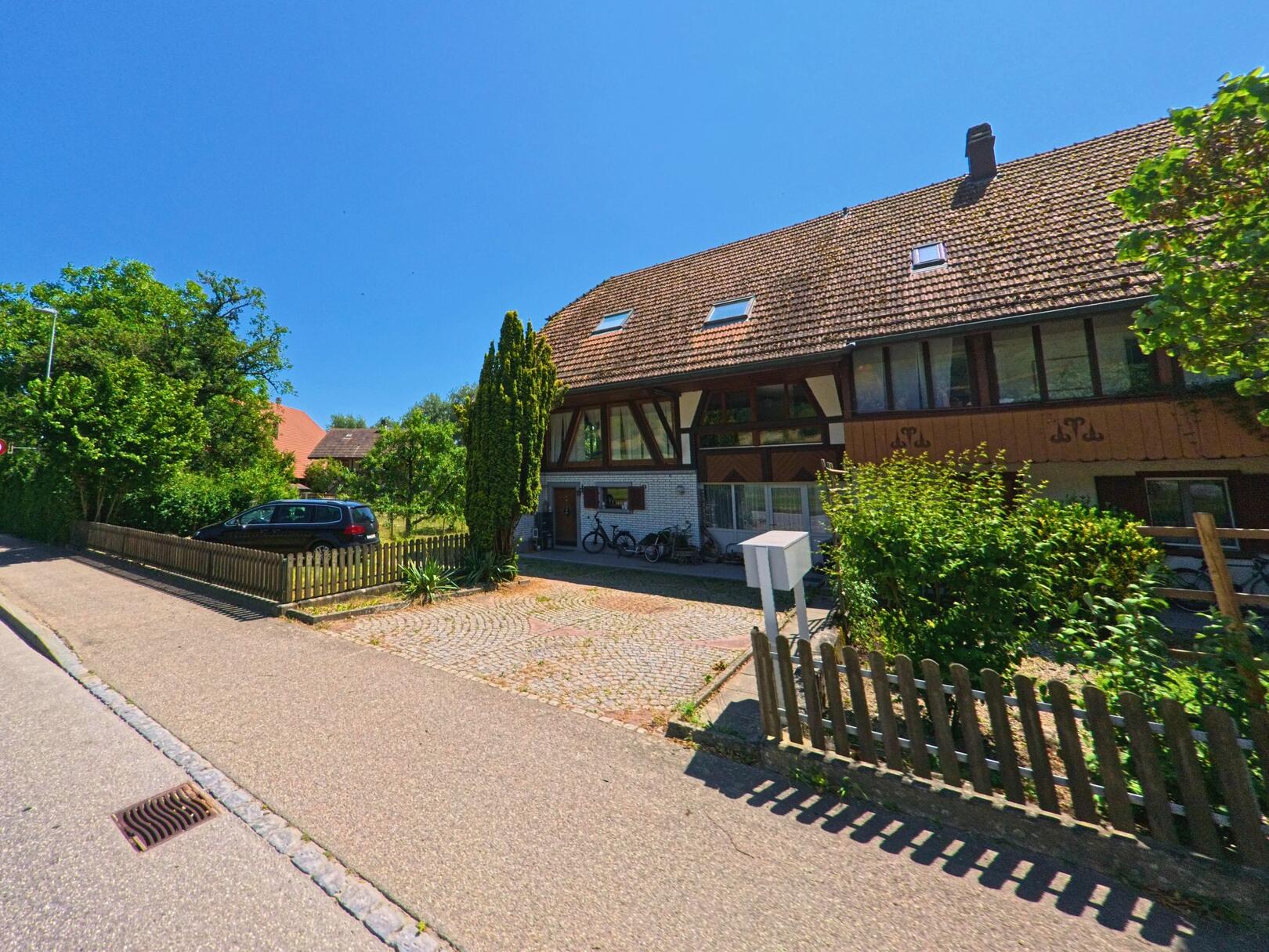 In Dotzigen (3293, Kanton Bern) zeigt sich ein traditionelles Wohnhaus mit Satteldach und sichtbarem Fachwerk, eingerahmt von Vorgarten, Kiesfläche und Holzlattenzaun.