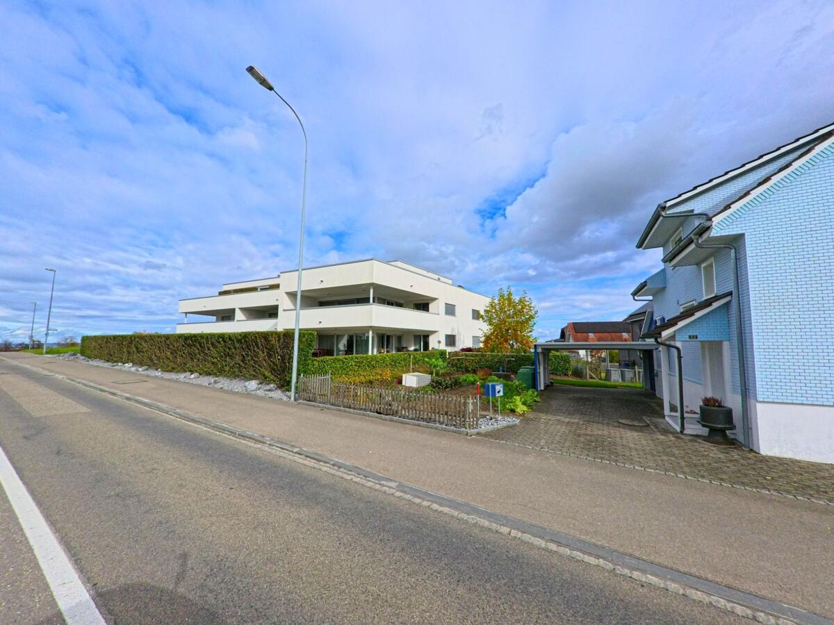 Ein modernes, weisses Gebäude mit grossen Fensterflächen steht in Dozwil (8582, Kanton Thurgau), flankiert von einer Hecke und einem angrenzenden Wohnhaus mit Carport.