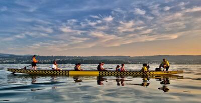 Ein Drachenboot mit mehreren Paddlerinnen und Paddlern gleitet bei ruhigem Wasser über den Zürichsee. Die Abendstimmung mit Spiegelungen auf dem See vermittelt Teamgeist, Ausdauer und die besondere Atmosphäre des Drachenbootsports.