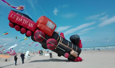 Am Strand fern von Zofingen (4800, Kanton Aargau) steigt ein grosser Lokomotiv-Drachen zwischen vielen bunten Figuren in den Himmel, waehrend Menschen im Sand stehen.
