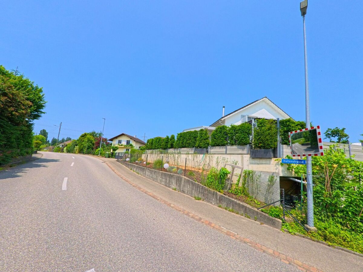 Die Haltenstrasse in Drei Höfe (4558, Kanton Solothurn) windet sich durch ein Wohnquartier mit gepflegten Gärten und einem Verkehrsspiegel an der Strassenecke.