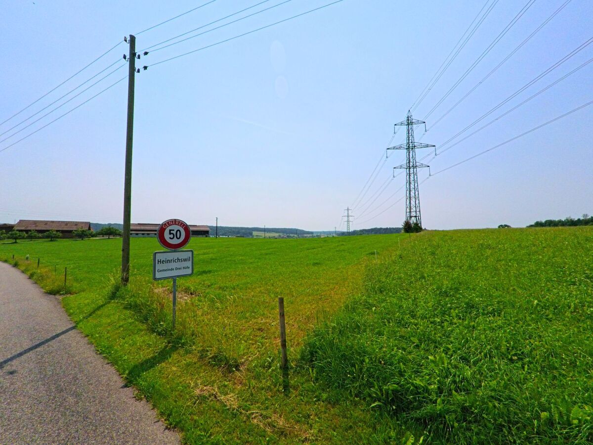 Ein Ortsschild markiert den Beginn von Heinrichswil in der Gemeinde Drei Höfe (4558, Kanton Solothurn), flankiert von weiten Feldern und einer Hochspannungsleitung.