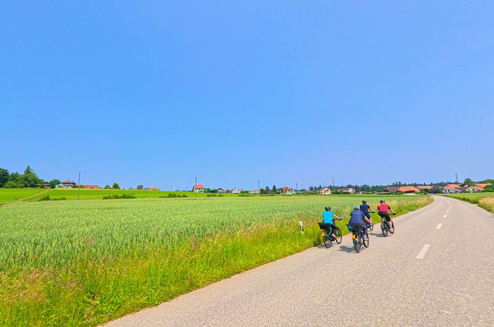 Bei strahlendem Sonnenschein fahren mehrere Personen auf Fahrrädern durch die offene Landschaft von Drei Höfe (4558, Kanton Solothurn), vorbei an Feldern und verstreuten Häusern.