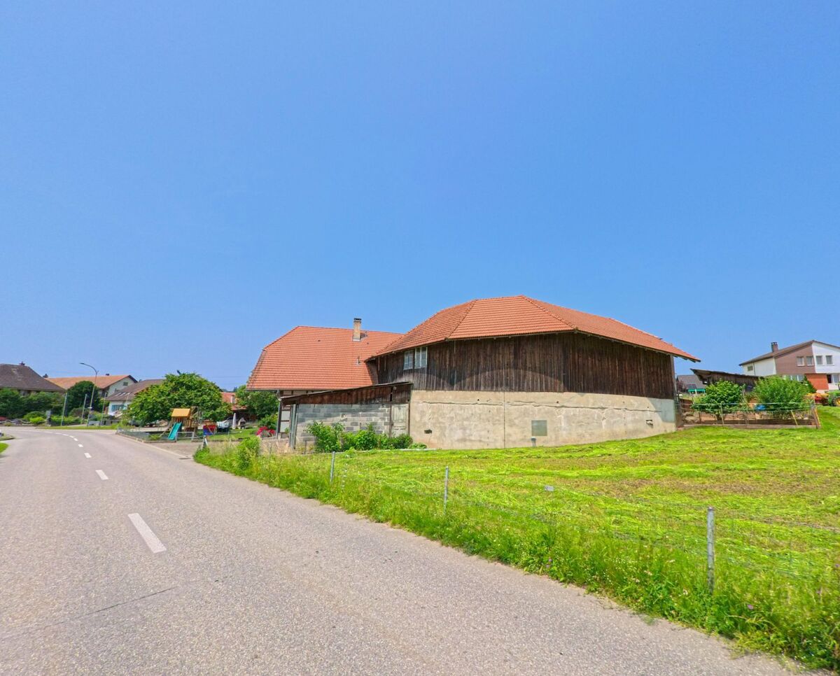 Ein altes Wirtschaftsgebäude mit rotem Ziegeldach steht in Drei Höfe (4558, Kanton Solothurn) am Rand einer Dorfstrasse, umgeben von Wiesen und Einfamilienhäusern.