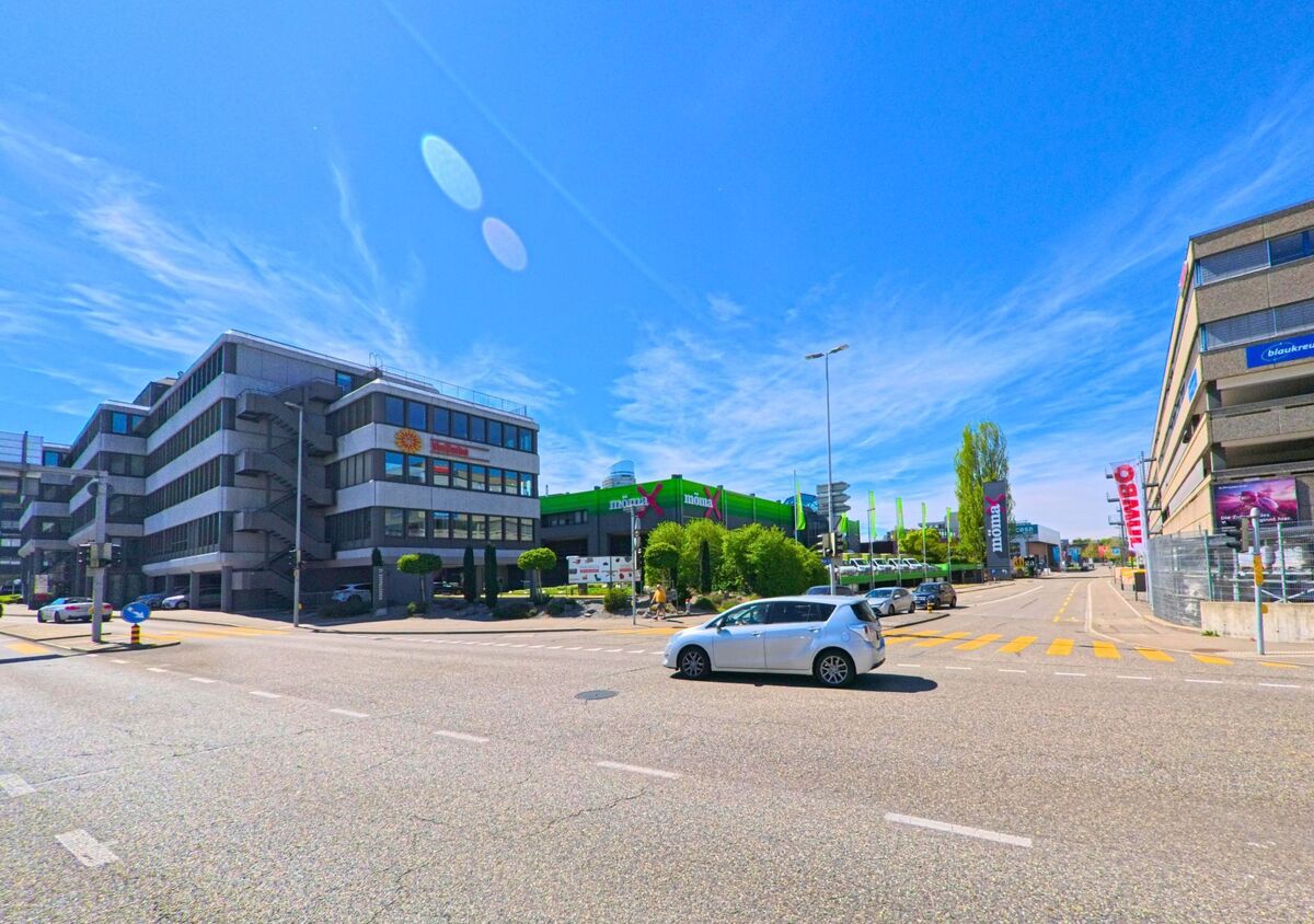 In [ORT], PLZ [PLZ], im [KANTON] verläuft eine breite Strasse durch ein Gewerbegebiet mit modernen Bürogebäuden, Geschäften und einem auffällig grünen Einkaufszentrum.