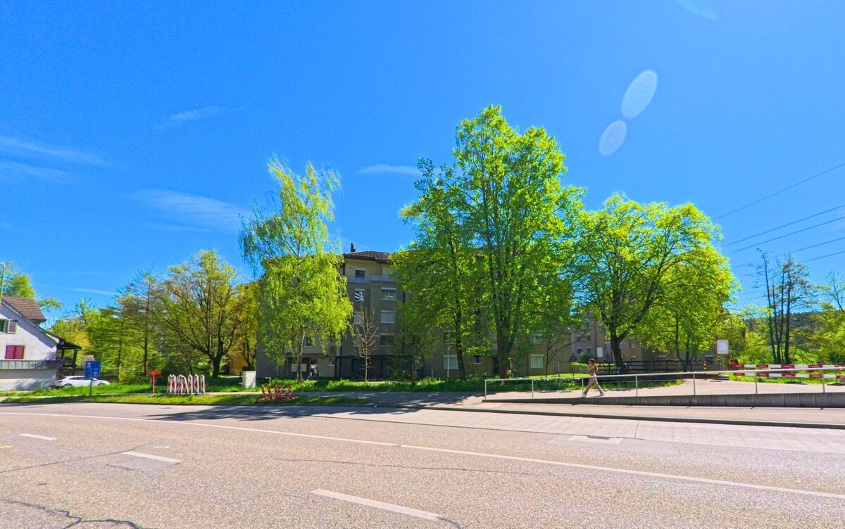 In [ORT], PLZ [PLZ], im [KANTON] stehen Wohnhäuser hinter dichtem Grün entlang einer mehrspurigen Strasse, die von Fussgängern und Radfahrern genutzt wird.