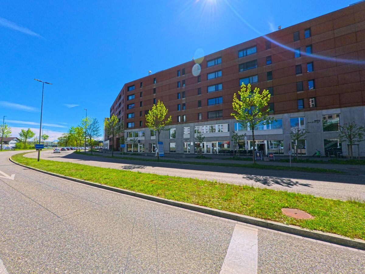 In [ORT], PLZ [PLZ], im [KANTON] erstreckt sich ein modernes Wohn- und Geschäftsgebäude entlang einer breiten Strasse mit junger Baumallee.