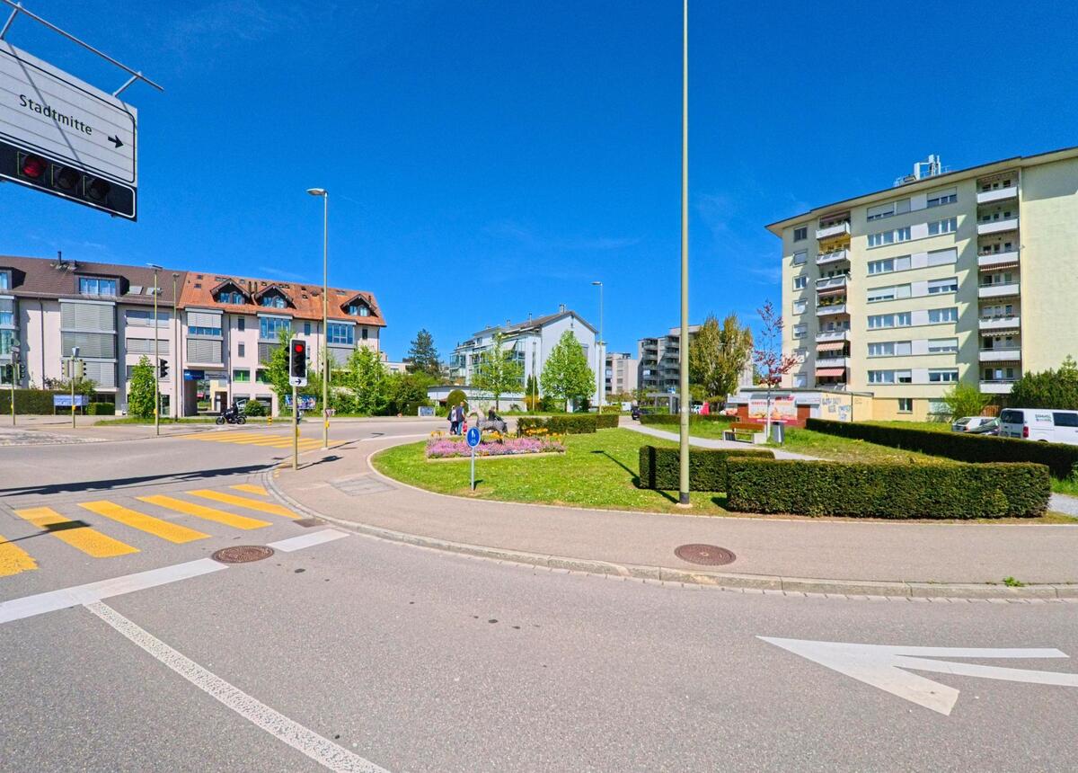 In [ORT], PLZ [PLZ], im [KANTON] kreuzen sich mehrere Strassen an einem Kreisverkehr mit Blumenbeet und Hinweisschild zur Stadtmitte, umgeben von Wohnhäusern.