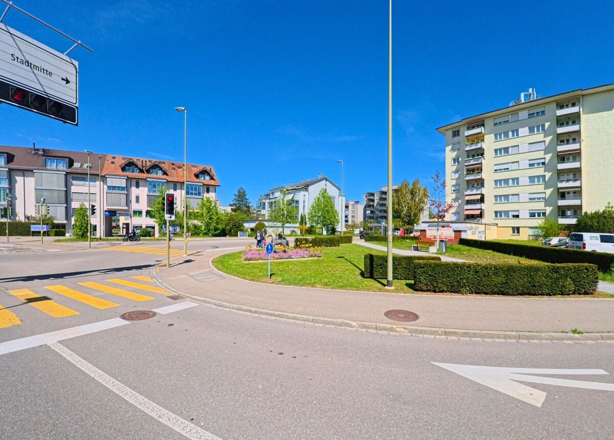 In [ORT], PLZ [PLZ], im [KANTON] kreuzen sich mehrere Strassen an einem Kreisverkehr mit Blumenbeet und Hinweisschild zur Stadtmitte, umgeben von Wohnhäusern.