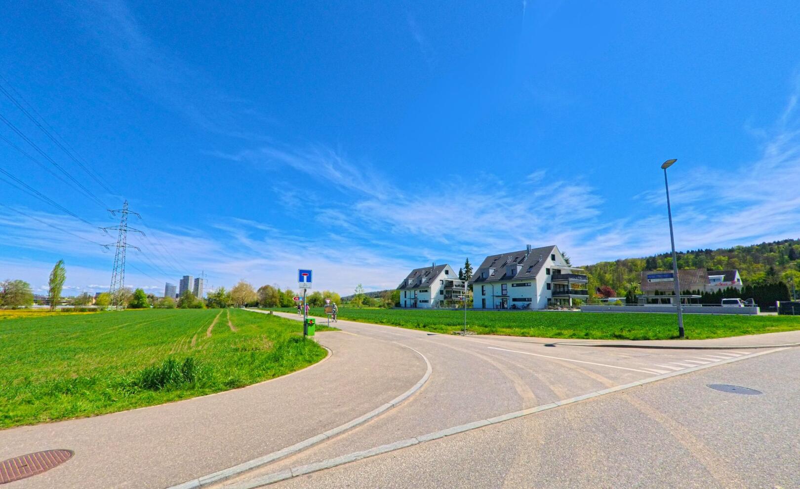 Am Siedlungsrand von Dübendorf (8600, Kanton Zürich) trifft eine asphaltierte Strasse auf Felder, während moderne Mehrfamilienhäuser vor bewaldeten Hügeln stehen.