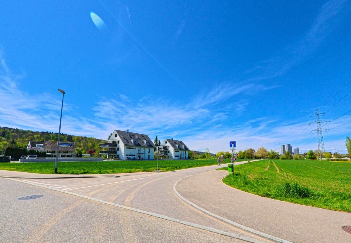 Am Rand von [ORT], PLZ [PLZ], im [KANTON] stehen moderne Mehrfamilienhäuser zwischen Wiesen und Feldern, im Hintergrund sind Hochspannungsmasten und hohe Gebäude zu sehen.