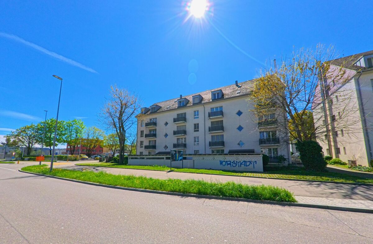 In [ORT], PLZ [PLZ], im [KANTON] steht ein mehrstöckiges Wohnhaus an einer breiten Strasse, im Vordergrund ist eine Mauer mit dem Schriftzug „Vorstadt“ zu sehen.