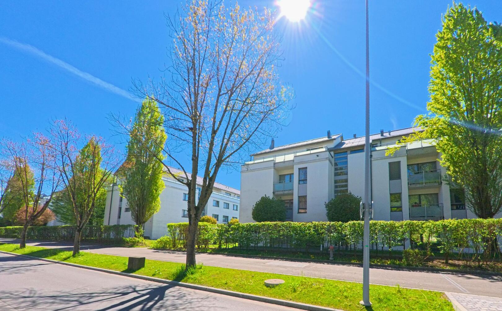 In [ORT], PLZ [PLZ], im [KANTON] stehen moderne Wohnhäuser entlang einer begrünten Strasse, durchflutet vom Sonnenlicht eines klaren Frühlingstages.